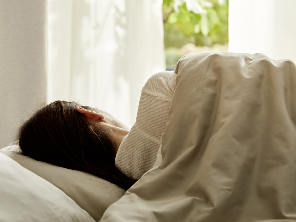 Eine Frau liegt im Bett mit ökologischen Schlafprodukten und Heimtextilien vor einem Fenster mit weißen Vorhängen
